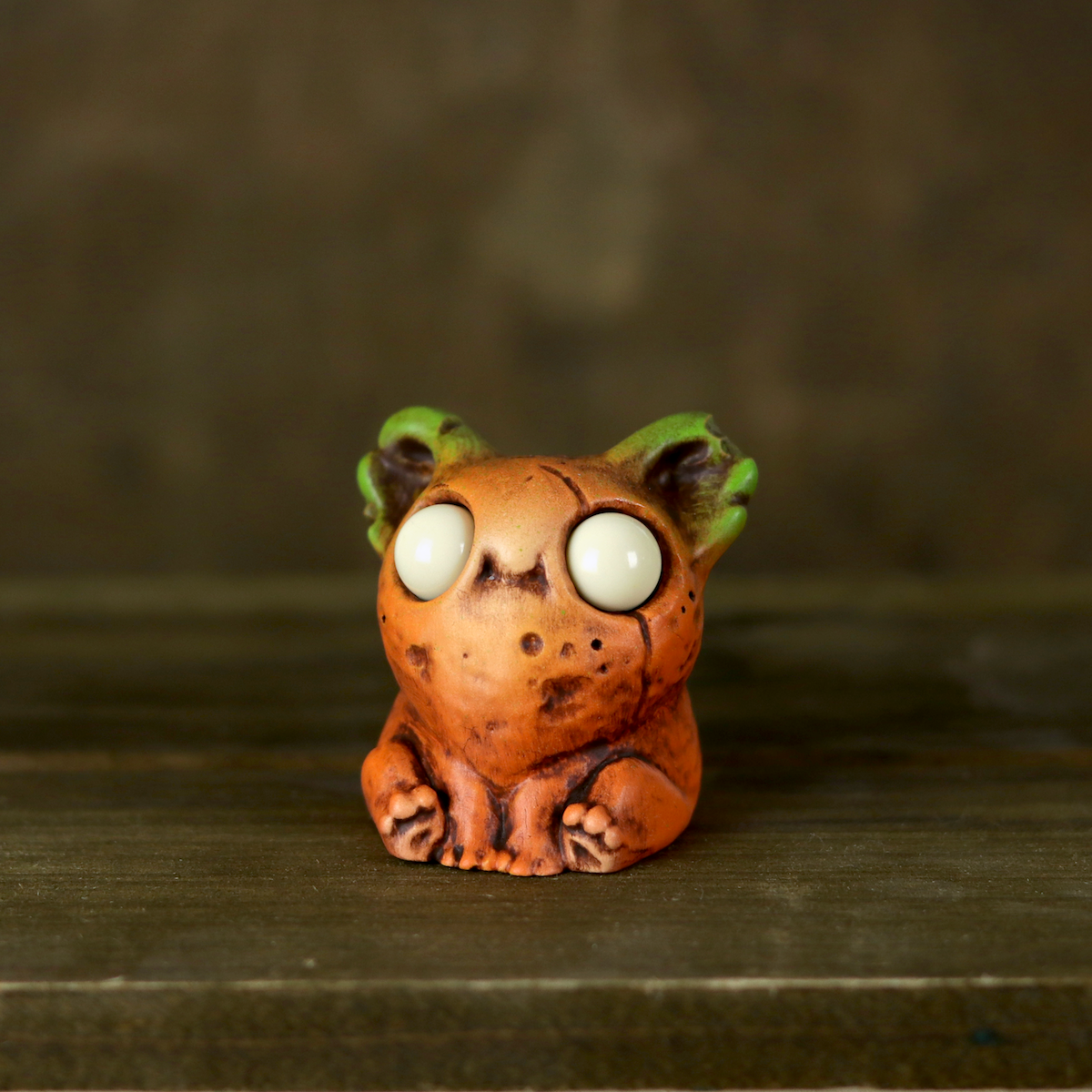 A Chris Ryniak Rotten Pumpkin Smidgen, a small orange clay creature with large white eyes and green-tipped ears, sits on a wooden surface against a dark background.
