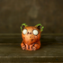 A Chris Ryniak Rotten Pumpkin Smidgen, a small orange clay creature with large white eyes and green-tipped ears, sits on a wooden surface against a dark background.