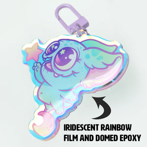 A whimsical charm featuring the "Candy Bat" creature from Bindlewood Shop, with a star and rainbow motif, enhanced by an iridescent rainbow film and coated with domed epoxy for a glossy finish.
