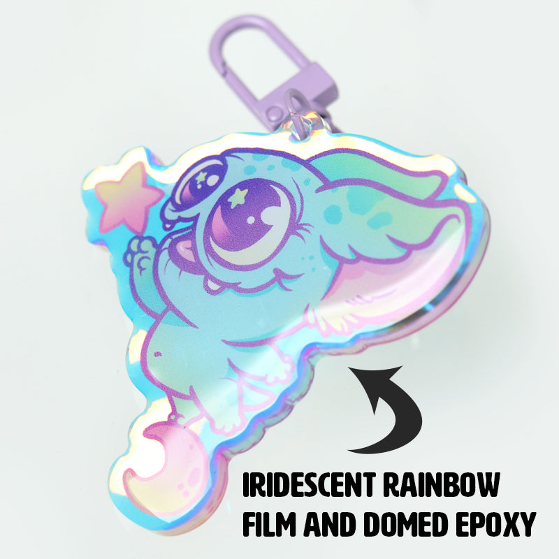 The Star Catcher Keychain by Bindlewood Shop features Chris Ryniak’s wide-eyed blue creature holding a pastel star, with iridescent rainbow film and a domed epoxy finish.