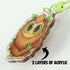 Close-up of a Lil' Reaper Keychain by Bindlewood Shop, featuring a cute brown creature with green leaves. A black arrow points to its two acrylic layers, labeled "2 LAYERS OF ACRYLIC" on a white background.