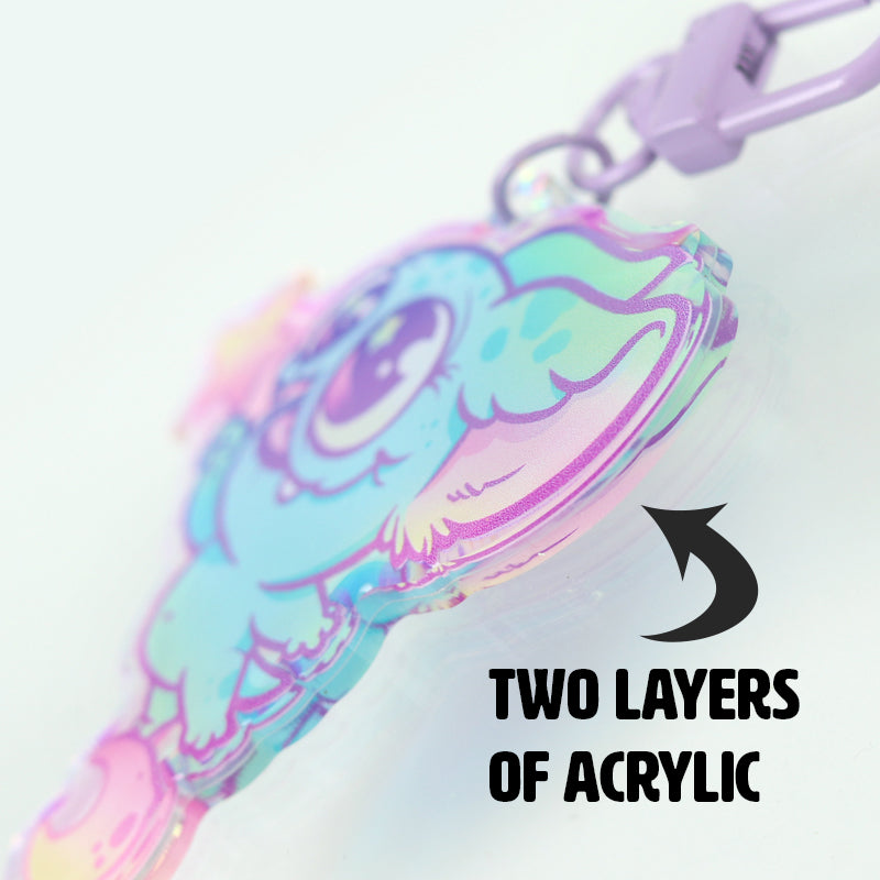 Close-up of the colorful, cartoon-style Flooficorn Keychain from Bindlewood Shop, shaped like an animal with big eyes and made from two thick layers of reflective acrylic. A black arrow and text say “TWO LAYERS OF ACRYLIC.”.