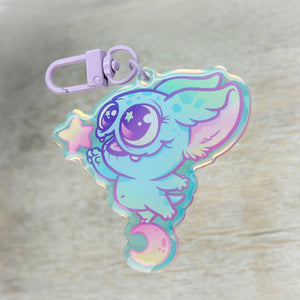 A whimsical keychain featuring the "Starcatcher" Rainbow Acrylic Charm designed by Bindlewood Shop, surrounded by stars and a crescent moon, in pastel colors, set against a wooden surface.