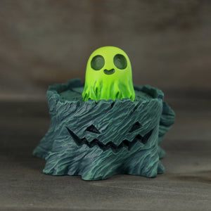 Introducing the "Spooky Stump Set" by Chris Ryniak: This delightful piece features a small, cheerful green ghost figurine emerging from a dark, rocky base adorned with a jack-o'-lantern design. Crafted in resin and hand-painted, this whimsical addition perfectly captures the spooky yet playful Halloween spirit against its gray backdrop.