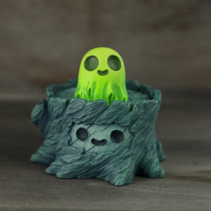 The Spooky Stump Set by Chris Ryniak showcases a small, green ghost with a cheerful expression nestled inside a carved tree stump. Both elements boast simple, cartoon-like features and are meticulously crafted from handpainted cast resin, set against a plain, dark textured background.