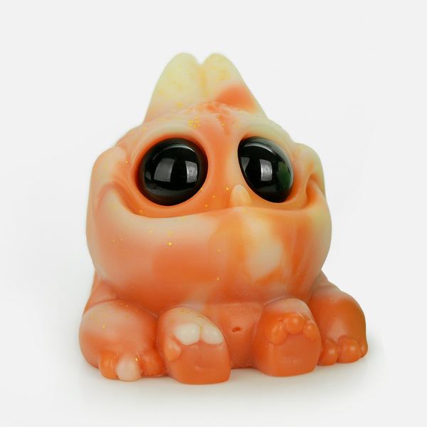 The Chris Ryniak Thai Tea Tank is a small, chonky, orange and cream-colored creature with big glossy black eyes, chubby cheeks, and tiny paws, posed cutely on a white background—just like a signature Ryniak resin figure.