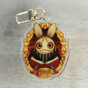 An illustrated keychain featuring a whimsical creature with rabbit ears reading a book entitled "Thimblestump Hollow," designed by Chris Ryniak and produced by Bindlewood Shop.