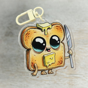 A cute Bindlewood Shop "Toast Boy" Acrylic Charm of a piece of toast with big eyes, holding a knife and a slice of cheese, with a little pat of butter melting on its head.
