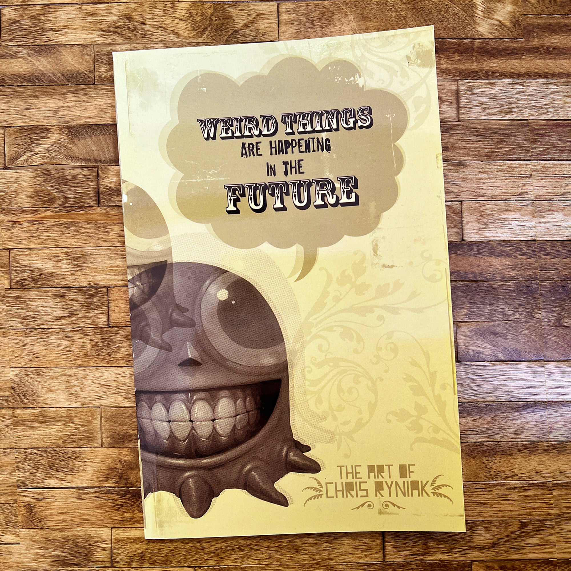 A yellow artbook, "Weird Things are Happening in the Future" by Bindlewood Shop, rests on a wooden surface. Its cover displays a grinning cartoon creature amid whimsical decorative swirls.