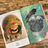 An open copy of "Weird Things are Happening in the Future" by Bindlewood Shop rests on a wooden table, showing pages with a round, green, human-faced creature with sharp teeth and a pug topped with a black crow against a green background.