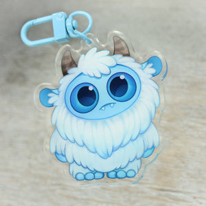 A cute blue and white Yeti charm keychain with big eyes and horns on a wooden surface by Bindlewood Shop.