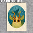A cute cartoon creature with big brown eyes, green fish scales, horns, and green ears sits in an oval frame. "Capricorn" tops this Zodiac Prints by Amanda Louise Spayd, available from Bindlewood Shop—perfect for any art collection.