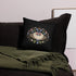 The Moth Magic Throw Pillow from Bindlewood Shop sits on a dark sofa next to a green blanket, with a framed picture propped against a light wall in the background.