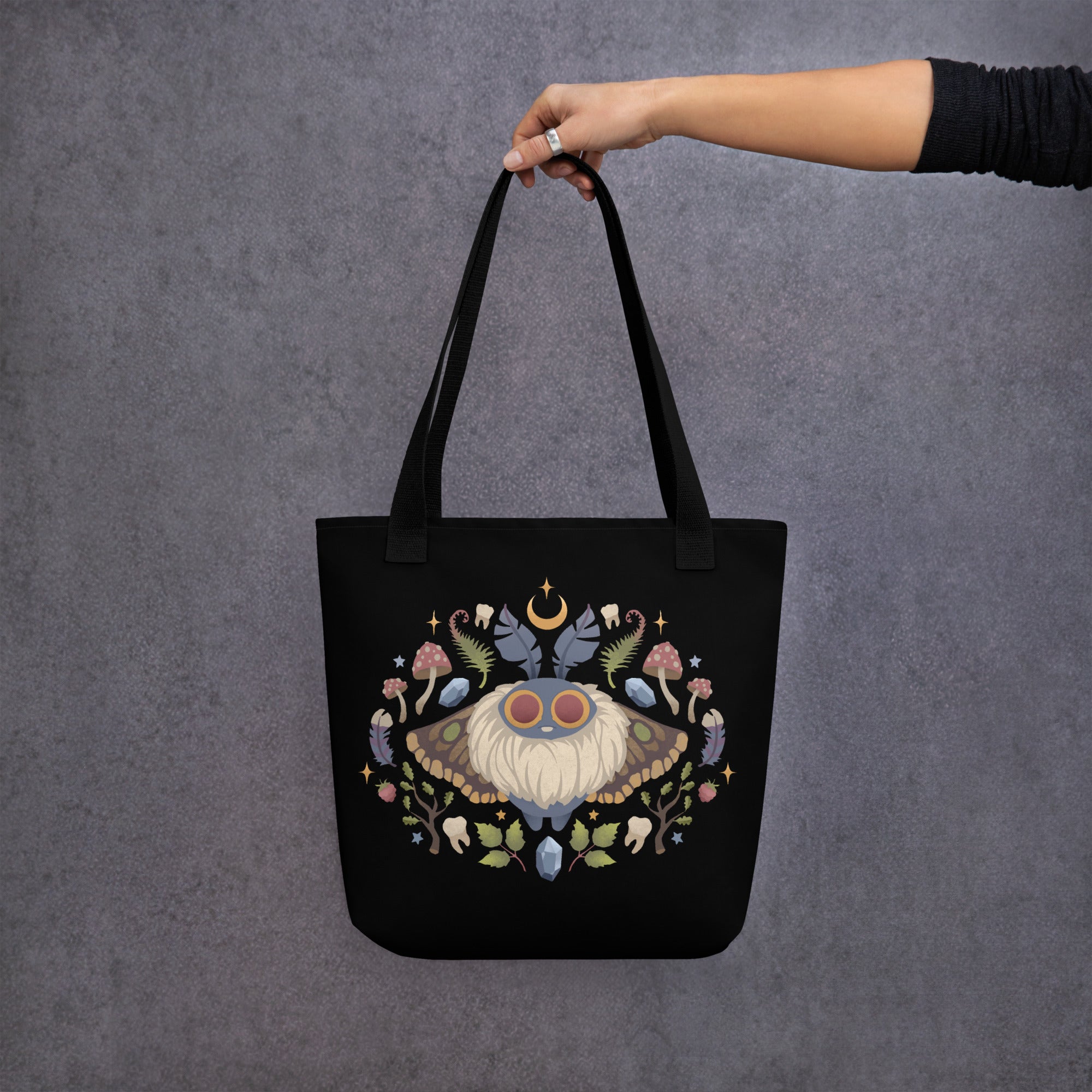 A hand holds Bindlewood Shop's Moth Magic Tote, featuring a colorful moth with flowers, foraged mushrooms, and celestial accents on a gray textured background.