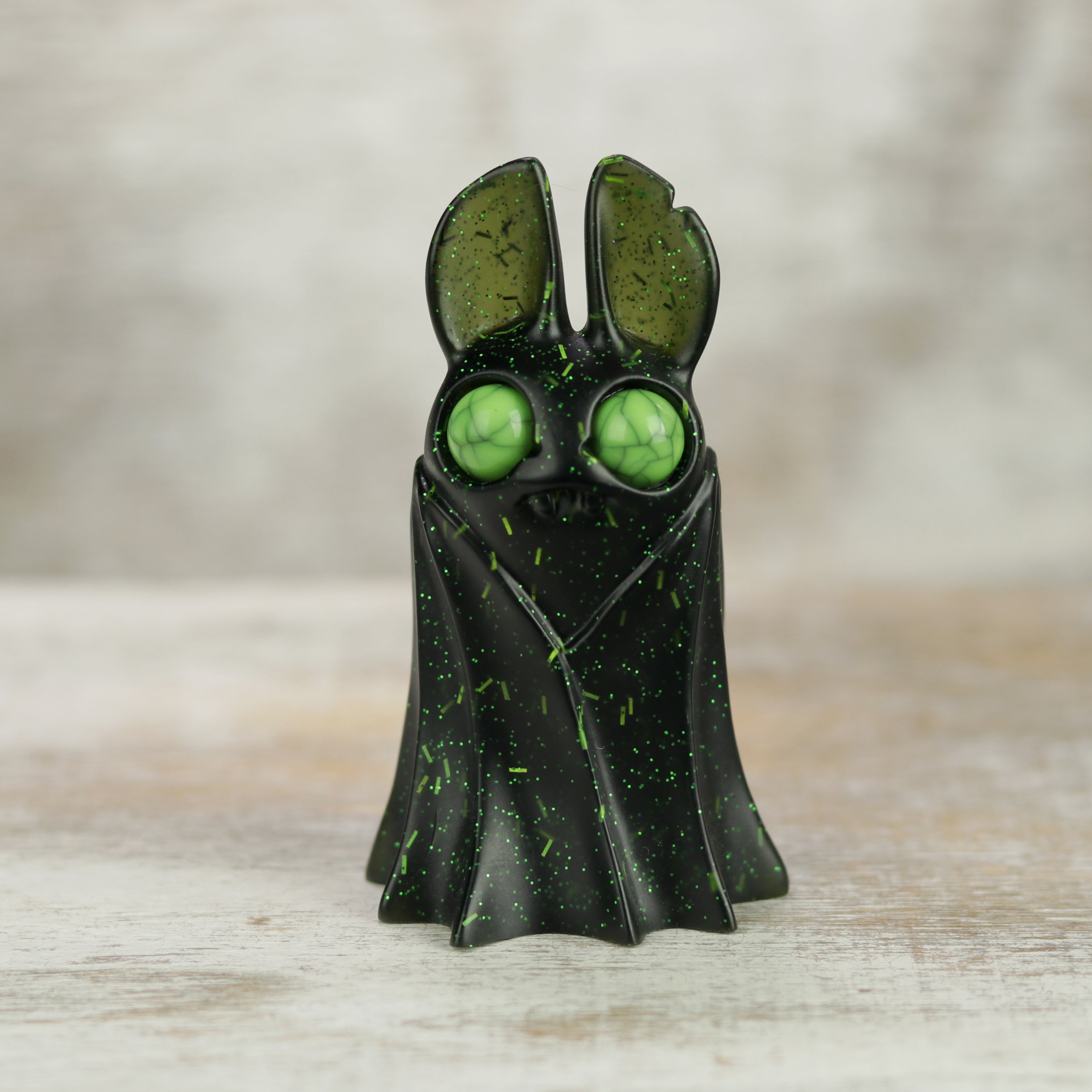 A small, black Holiday Belfry (Green) resin figure by Bindlewood Shop stands on a light wooden surface; it features green speckles, large round green eyes, and tall ears against a blurred neutral background.