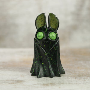 A small, black Holiday Belfry (Green) resin figure by Bindlewood Shop stands on a light wooden surface; it features green speckles, large round green eyes, and tall ears against a blurred neutral background.