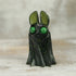 A small black resin bat figure with large green eyes and ears, accented by green speckles, stands on a wooden surface—a charming Holiday Belfry (Green) by Bindlewood Shop.