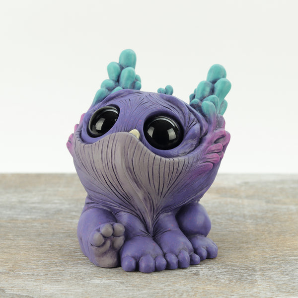 Dewlap by Chris Ryniak: a small, whimsical purple creature figurine with large black eyes, teal horns, wrinkled texture, and tiny feet sits on a wooden surface against a light background.