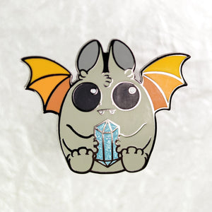 Illustration of a cute, cartoon-style bat with oversized eyes and small fangs, holding a sparkling blue crystal. The bat has gray fur, orange wings, and is set against a plain white background featuring the Atticus Enamel Pin from Bindlewood Shop.