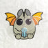 Illustration of a cute, cartoon-style bat with oversized eyes and small fangs, holding a sparkling blue crystal. The bat has gray fur, orange wings, and is set against a plain white background featuring the Atticus Enamel Pin from Bindlewood Shop.