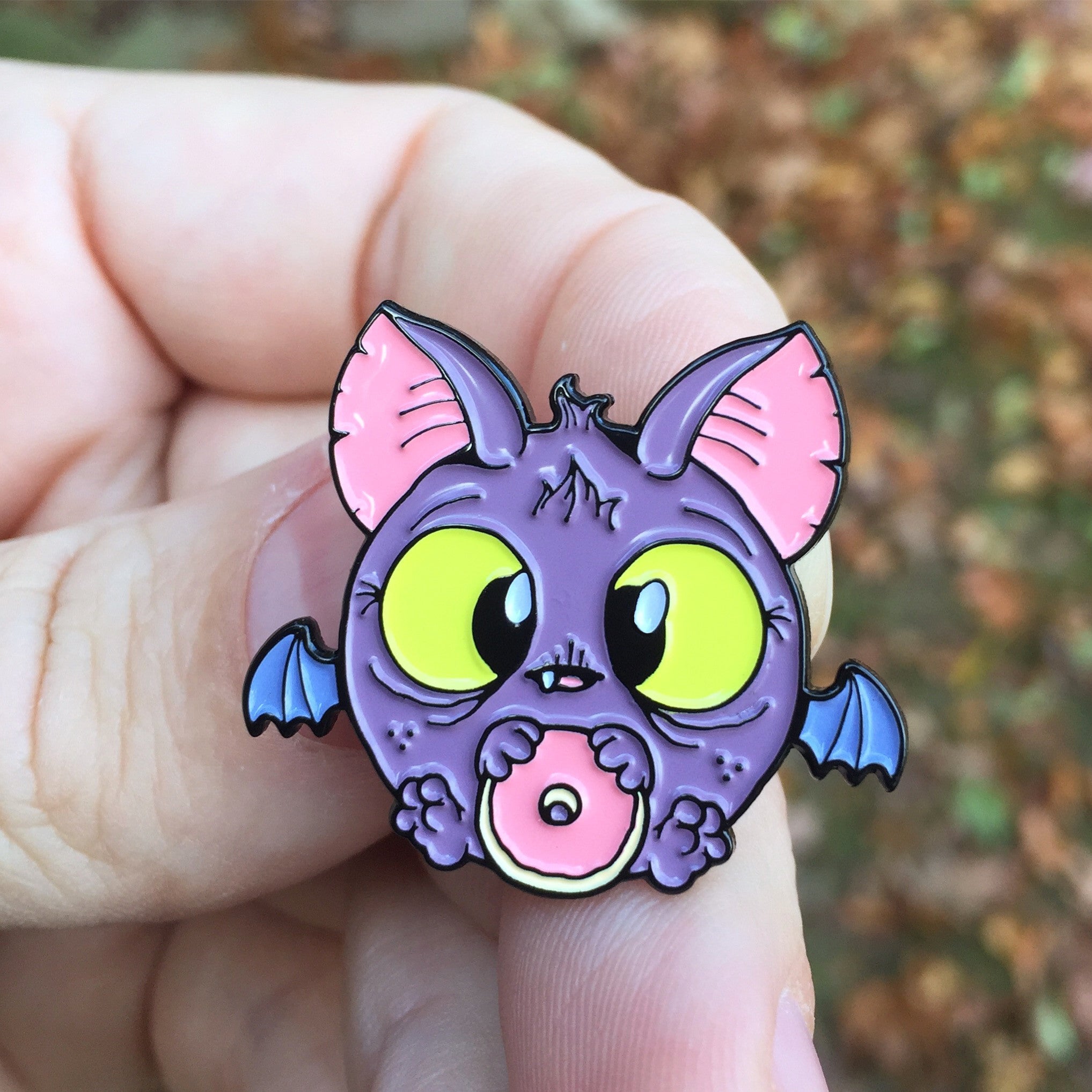 A hand holds the Bindlewood Shop Donut Bat Pin—an enameled metal pin of a chubby purple bat with yellow eyes, pink ears, and blue wings clutching a bitten pink frosted donut. Blurred foliage fills the background.