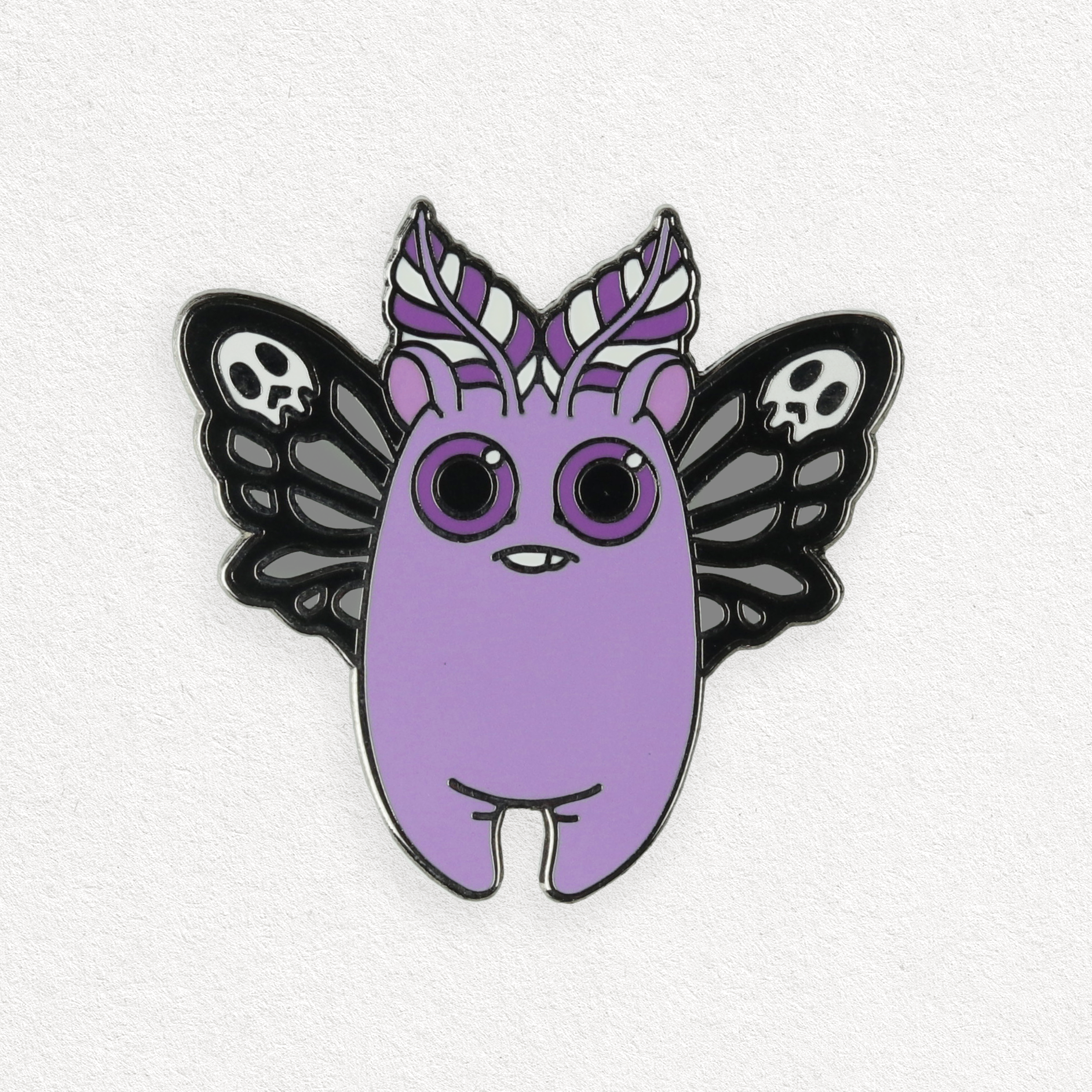 A whimsical hard enamel pin featuring a Death's Head Mothbunny Pin from Bindlewood Shop, showcasing big round eyes and feathery wings accentuated with skull motifs.