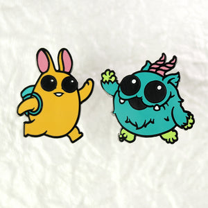 Two colorful High Five Enamel Pin Set from Bindlewood Shop, on a white background: a yellow rabbit-like figure with large eyes and headphones, and a blue, fluffy monster with a unicorn horn and a leaf.