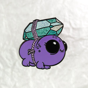 A cute, purple enamel pin depicting a Gemscuffler from Thimblestump Hollow with a large, sparkly green crystal growing on its back, braided detailing, and big, round eyes by Bindlewood Shop.