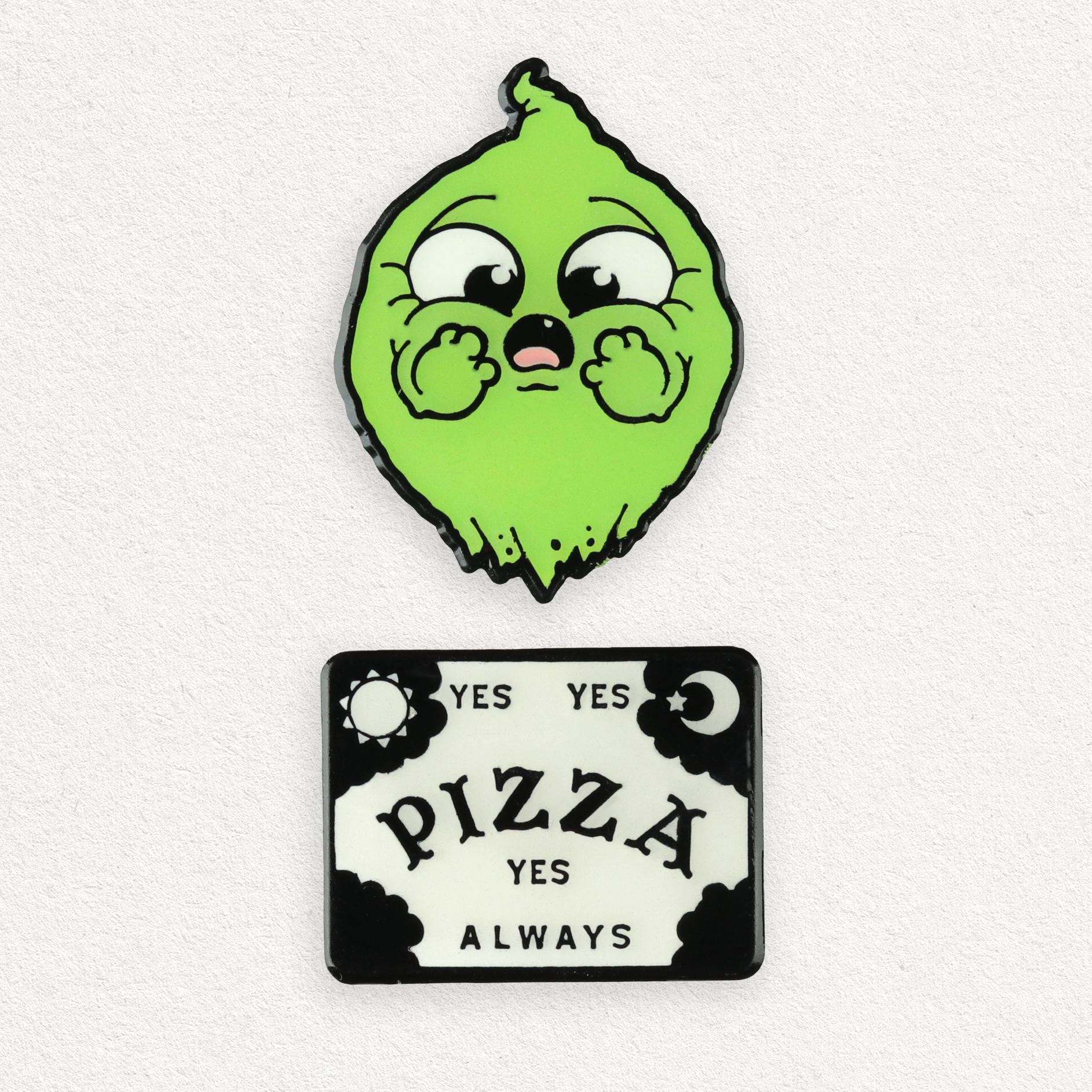 The OMGhost Pin Set by Bindlewood Shop features a cute green cartoon character pin with big eyes and a black and white “YES YES PIZZA YES ALWAYS” spirit board-style pin with moon and sun corners, shown on a plain light background.