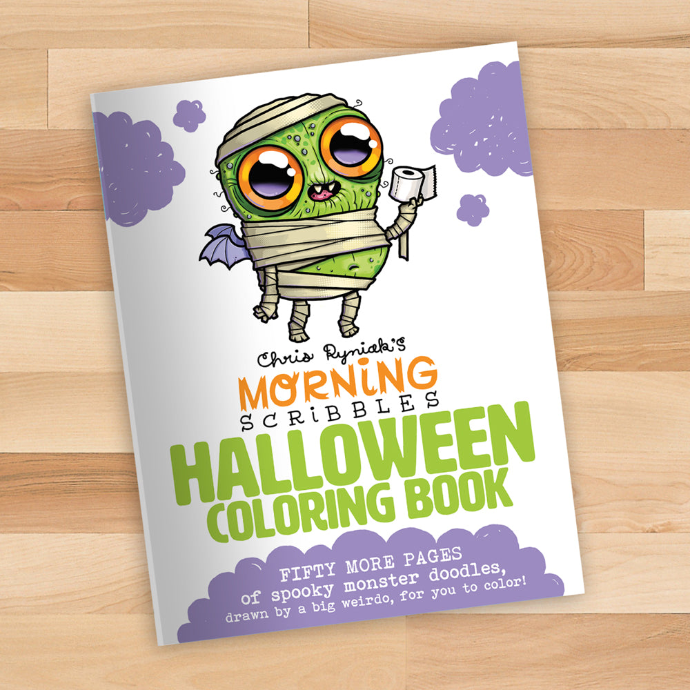The cover of the "Morning Scribbles Coloring Book - Halloween Edition" from Bindlewood Shop lies on a wooden surface, featuring a cute cartoon mummy with large, expressive eyes, bat wings, and purple clouds. An exclusive pre-order postcard is included, along with fifty more pages of spooky monster doodles.