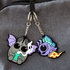 Two cute PVC rubber keychains hang side by side: one is a bat holding a skull, and the other is the Mothpup Keychain by Bindlewood Shop, featuring a purple insect-like creature with a blue fin and polka-dotted tail.