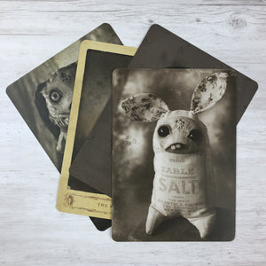 A set of three Dust Bunnies Cabinet Card photographs with illustrations of whimsical creatures, each blending an animal with an inanimate object, displayed on a wooden surface by Bindlewood Shop.
