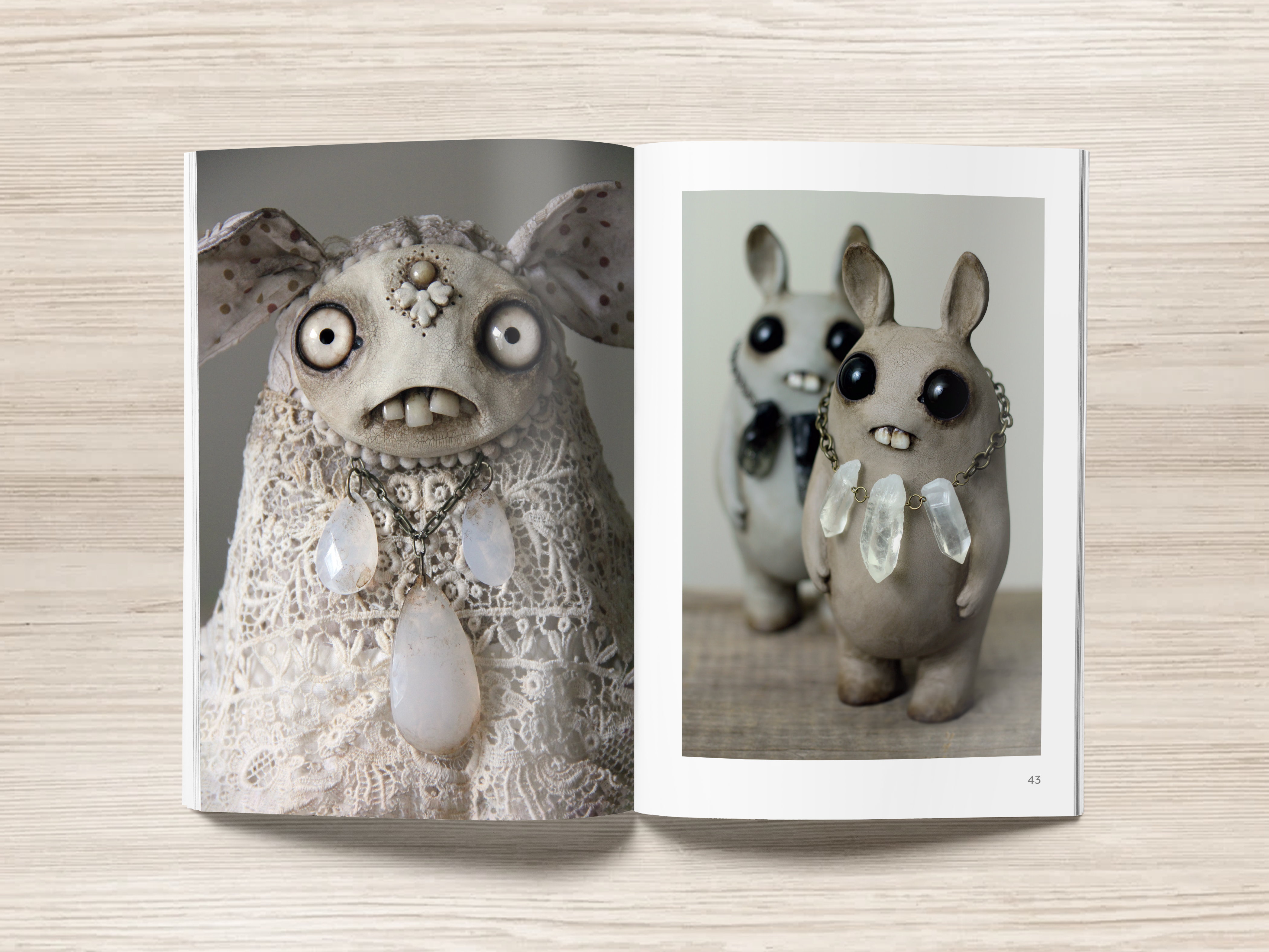 On a wooden surface lies an open softcover copy of Bindlewood Shop's "Making Friends" book. The left page showcases a whimsical bunny dressed in lace adorned with large gemstone necklaces. The right page, reminiscent of an Amanda Louise Spayd creation, features two creatures with dark eyes and matching necklaces.