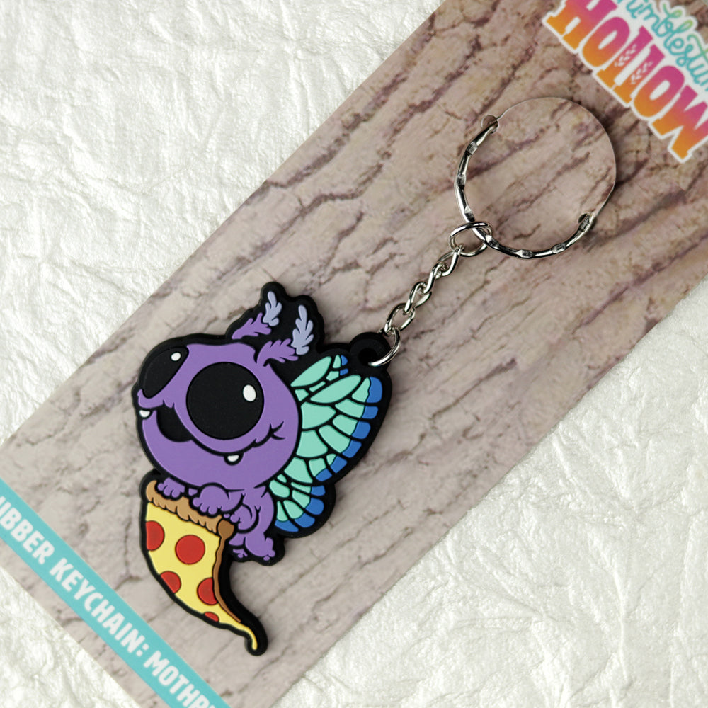 The Bindlewood Shop Mothpup Keychain features a cute purple moth with blue-green wings holding pizza, attached to a backing card. Perfect for Thimblestump Hollow fans!.
