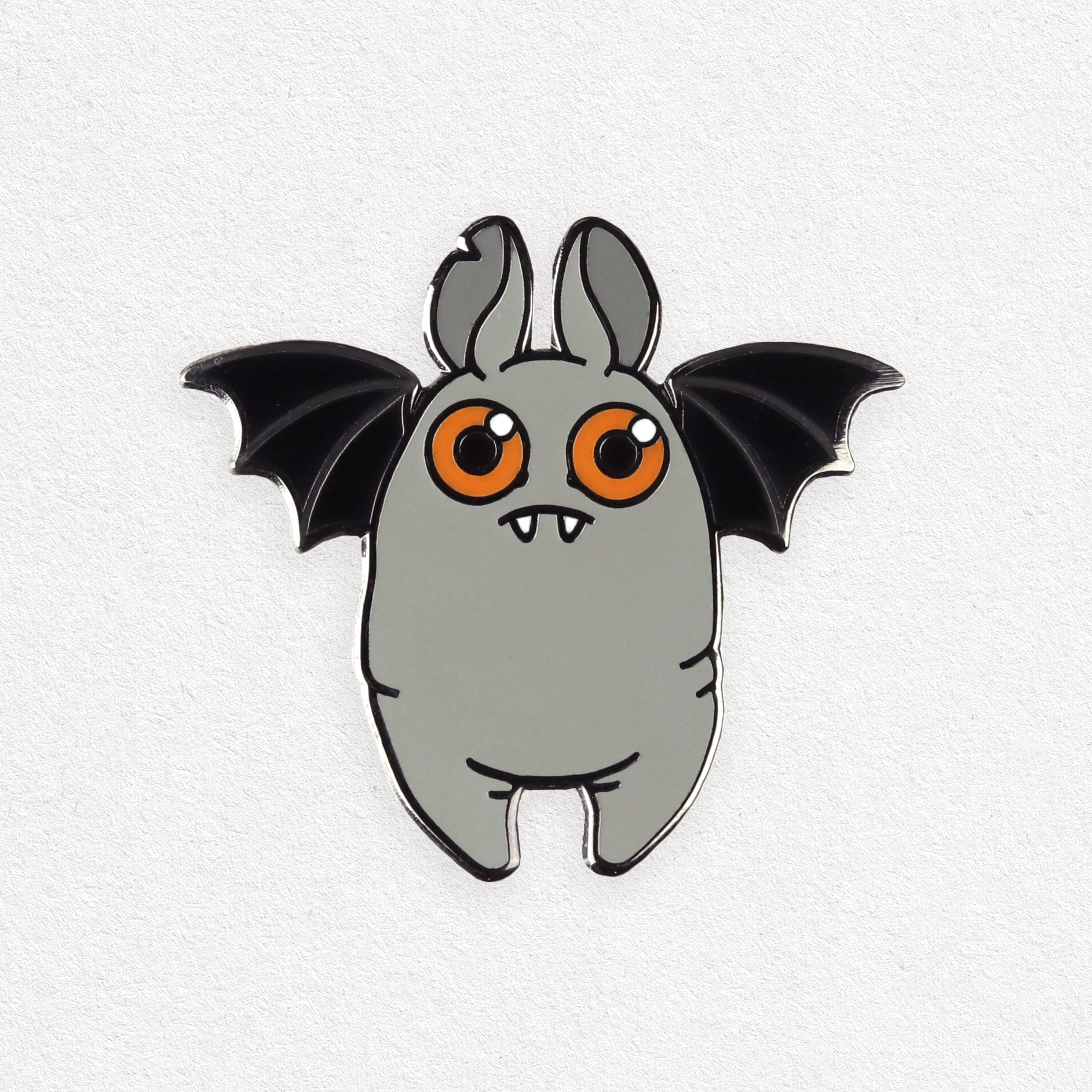 The Bindlewood Shop Orange-eyed Bat Pin is a cute, cartoon enamel pin featuring a chubby bat with round orange eyes, small fangs, upright ears, and black wings on a plain white background.
