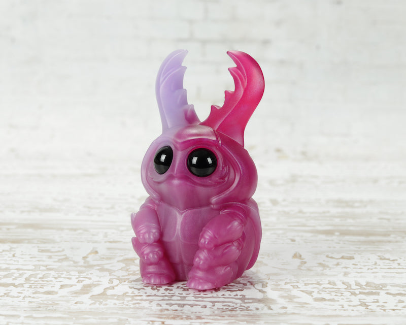 The MISFIT Sparkleberry Staggle by Bindlewood Shop is a small, purple-pink resin figure with large black eyes, stubby legs, and two curved translucent antennae sitting on a white textured wooden surface.