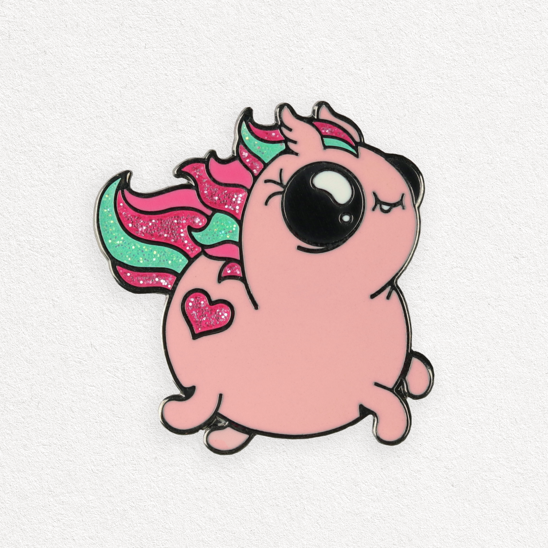 A cute enamel pin featuring a pink unicorn with a chubby body, large black eyes, and a small, smiling mouth. The Sparkle Pony Magic Pin from Bindlewood Shop showcases a colorful mane and tail with pink and green stripes, glittery sparkles, and a pink heart design on its side. The background is white.