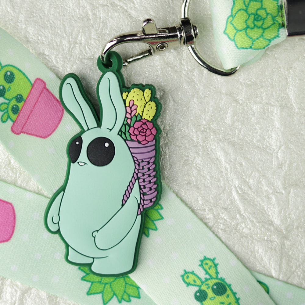 The Bindlewood Shop Succulent Lanyard features cacti and potted plants prints, and comes with a keychain of a green cartoon bunny carrying a basket of colorful succulents.