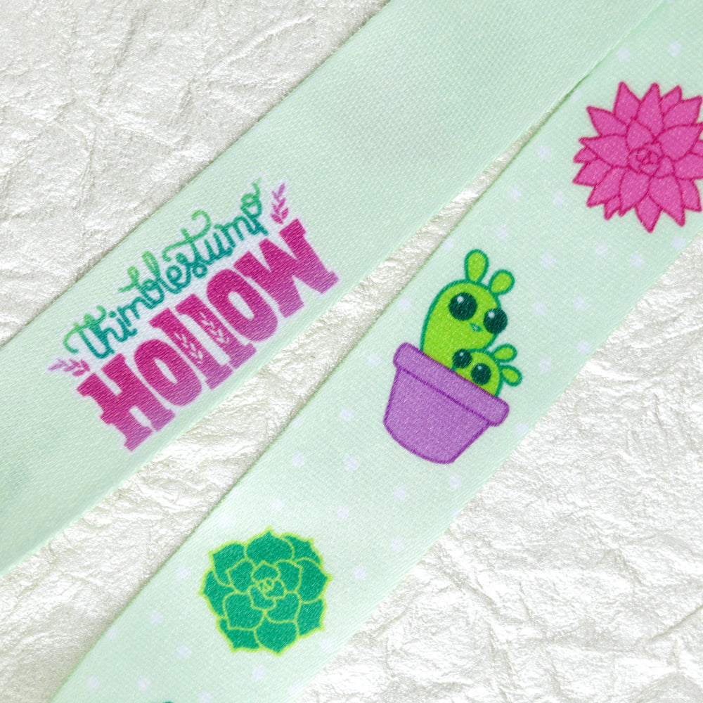 The Succulent Lanyard by Bindlewood Shop is light green with “Thistlebum Hollow” and features a pink flower, green succulent, and two cacti in a purple pot on a textured white background—perfect as a badge holder.