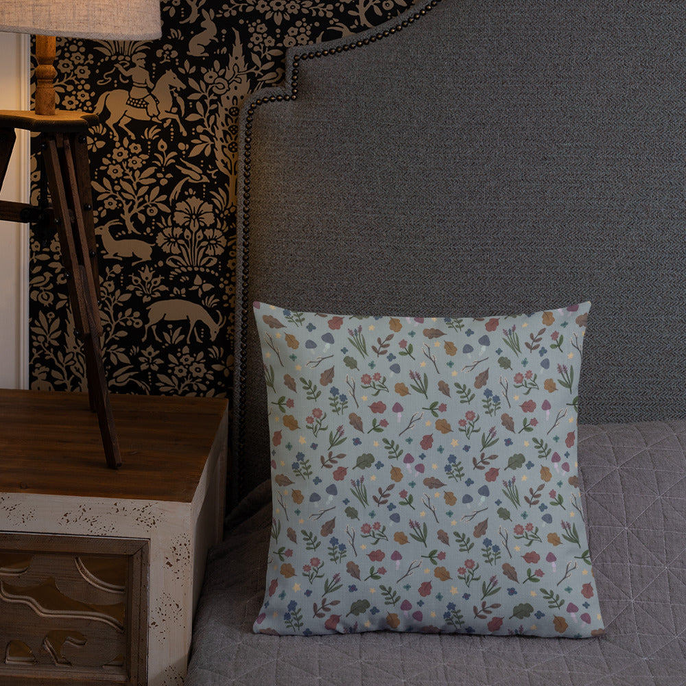 A Bindlewood Shop Twilight Sprite Throw Pillow with a floral pattern rests on a neatly made bed with a gray quilt. The headboard of the bed is upholstered with gray fabric. A wooden nightstand with a lamp, featuring nature-themed wallpaper in the background, complements the cottagecore interior design.