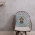 A metal wire chair with a cushioned seat features the charming Twilight Sprite Throw Pillow from Bindlewood Shop, adorned with an illustration of a cute bear wearing a dress, framed by a wreath of colorful flowers against a light blue background. The chair sits in a simply decorated room, evoking cottagecore interior design charm.