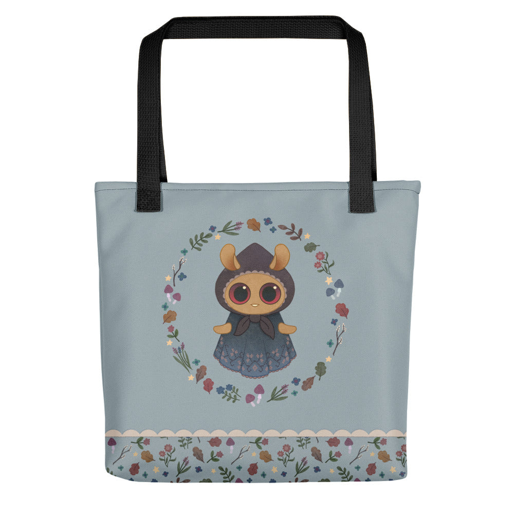 The Twilight Sprite Tote by Bindlewood Shop is a light blue, sturdy tote bag adorned with an illustration of an adorable character with big eyes, dressed in a cloak, and encircled by a floral design. Made from 100% spun polyester fabric, the bottom edge features a floral border on a white background. The handles are black and showcase the Bindlewood logo.