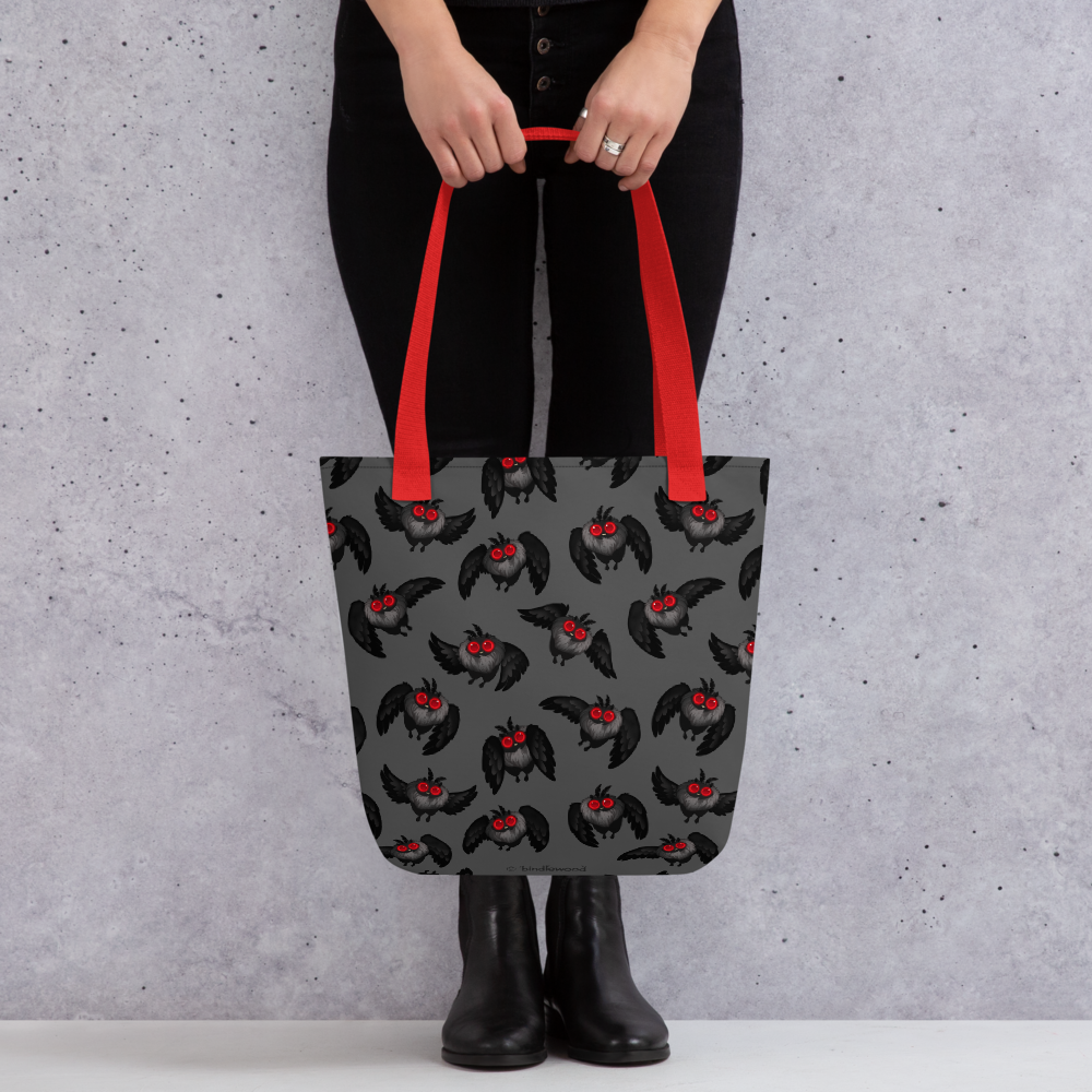 A person dressed in black pants and boots is holding a gray Mothman Cryptid Tote from Bindlewood Shop with red handles, featuring a pattern of black bats with red eyes and open wings. The durable fabric tote is perfect for carrying cryptid-hunting paraphernalia against a light gray, textured background.