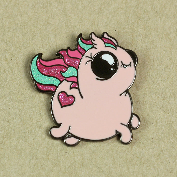 An enamel pin of a cute, chubby unicorn with a pink body, large black eyes, a tiny horn, and a multicolored mane and tail. It has a heart shape on its side and is set against a beige background. This Sparkle Pony Magic Pin from Bindlewood Shop brings an extra touch of whimsy to your collection.