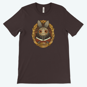 A brown "The Maker" Tee featuring a whimsical graphic of a cute yet slightly menacing character designed by Amanda Spayd, with large eyes, prominent teeth, and a bow tie, surrounded by ornate golden decorations. Available at Bindlewood Shop.