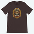 A brown "The Maker" Tee featuring a whimsical graphic of a cute yet slightly menacing character designed by Amanda Spayd, with large eyes, prominent teeth, and a bow tie, surrounded by ornate golden decorations. Available at Bindlewood Shop.
