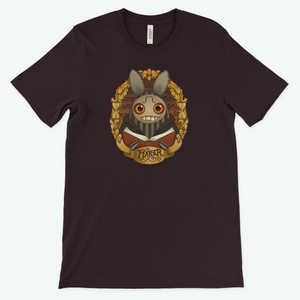 A "The Maker" Tee featuring a quirky illustration of a creature with large eyes, pointy ears, and a toothy grin, surrounded by ornate gold patterns and holding a heart with the word.