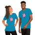 A woman and a man are smiling while wearing matching blue T-shirts featuring Chris Ryniak's colorful Flooficorn. The unicorn is predominantly pink with purple accents and is surrounded by small stars and sparkles. The woman has her hair styled in a bun, and the man has short hair. They are wearing the Floofy Unicorn Magic Tee from Bindlewood Shop.