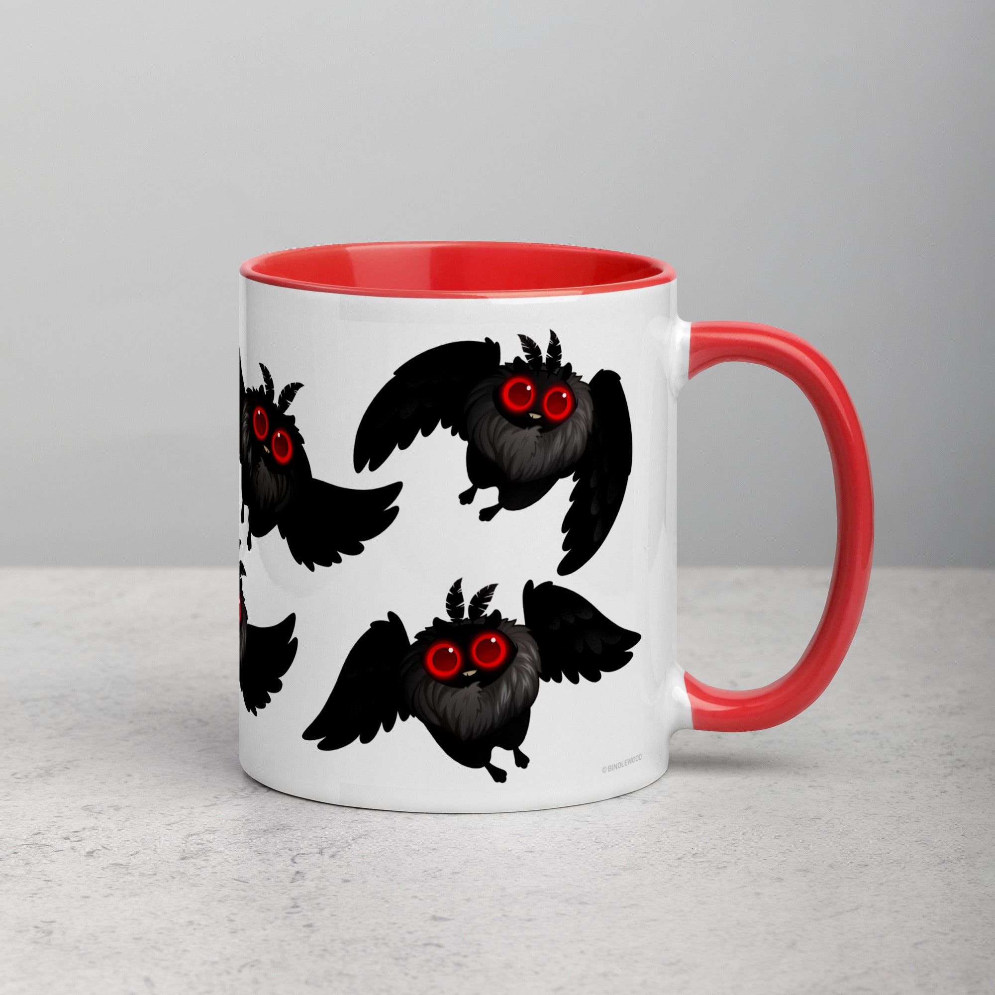 A white ceramic mug with a red interior and handle is shown on a light gray surface. The Mothman Cryptid Mug from Bindlewood Shop features a repeating design of cute black, fluffy creatures with bright red eyes and wings, reminiscent of Amanda Louise Spayd's whimsical style. The creatures appear to be in different flying poses.