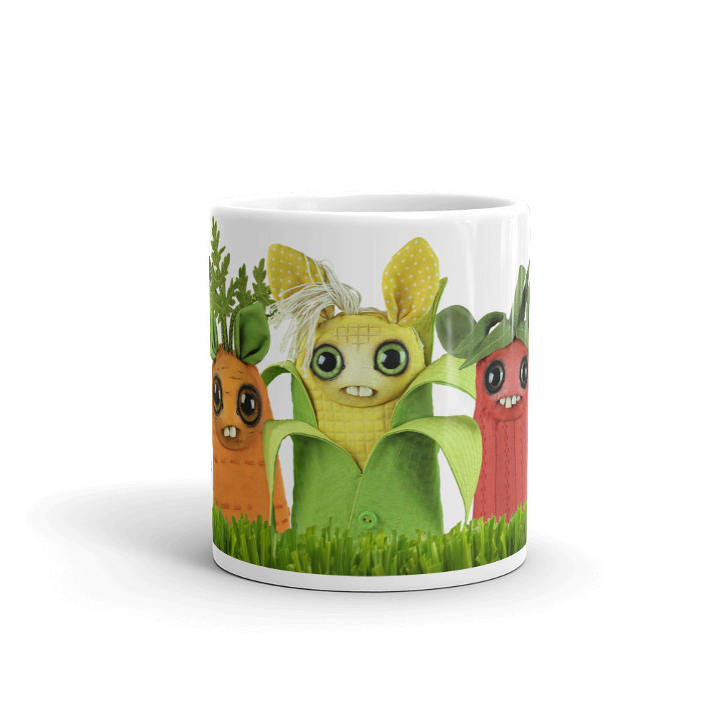 Introducing the Farm Market Dust Bunnies Mug by Bindlewood Shop: a microwave and dishwasher safe ceramic mug adorned with charming, anthropomorphic vegetable characters. Meet an orange carrot, a yellow corn with a bow, and a red radish—all featuring big eyes and cheerful smiles. The background at the bottom is decorated with green leaves and grassy details for an extra touch of whimsy.